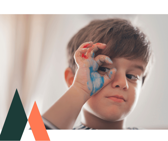 Child with their right hand covered in paint, making a circle gesture around one eye while smiling playfully.