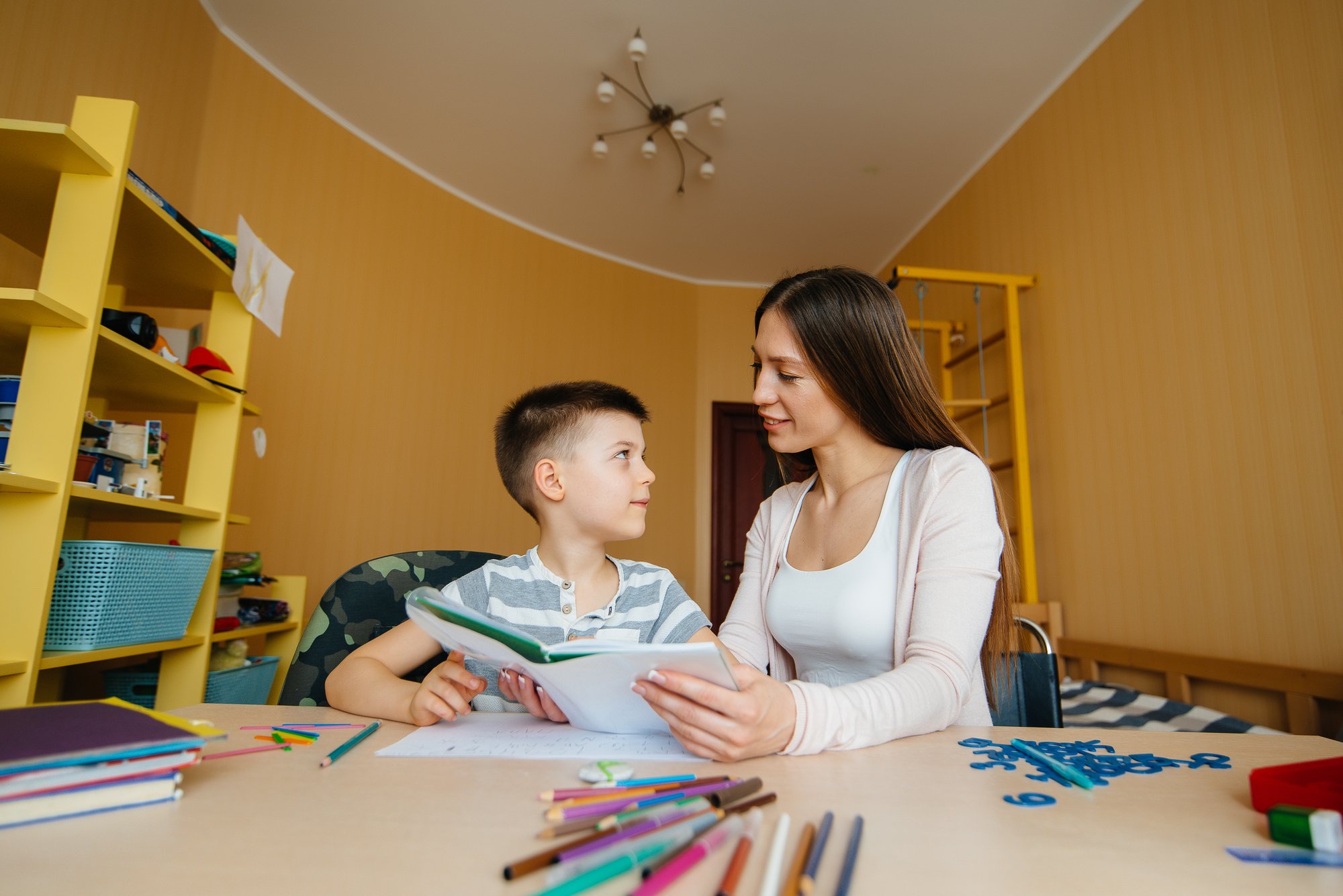 	Therapist and child facing each other in a bright yellow behaviour therapy room, each holding a sheet of paper and sharing a focused smile.
