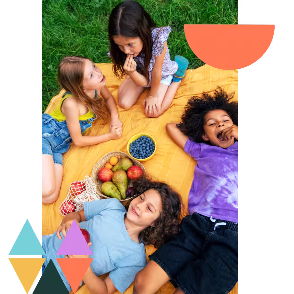Group of children lying on a picnic blanket outdoors, sharing fresh fruit together during a community activity